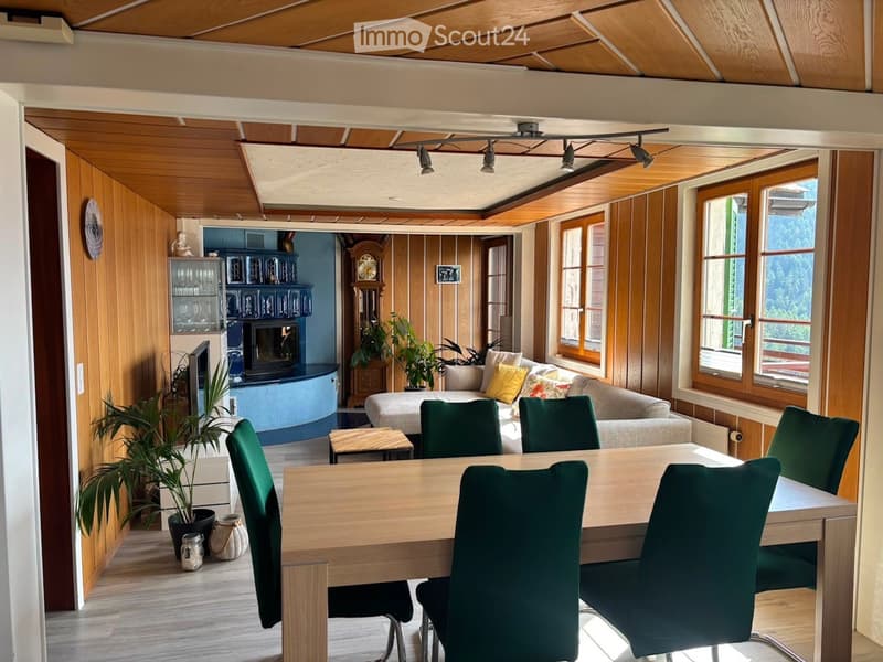 A spacious, well-lit living and dining area with wooden ceilings and walls, featuring a sofa, dining table with chairs, fireplace, clock, and potted plants.