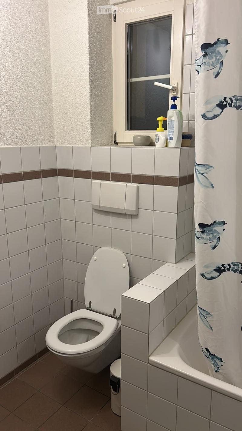 A small bathroom with white tiled walls, a toilet, a shower curtain, and a small window above the sink area.