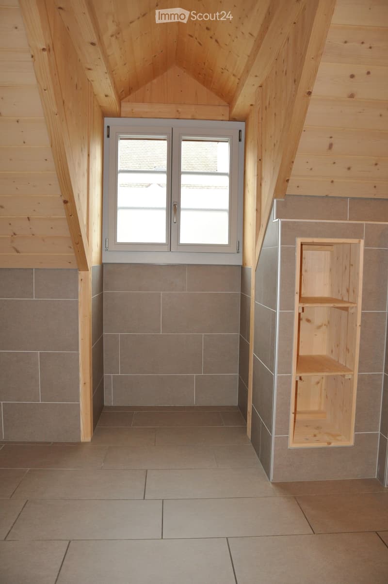 Empty attic space with wooden beams, tiled floor, and a small built-in wooden shelf unit.