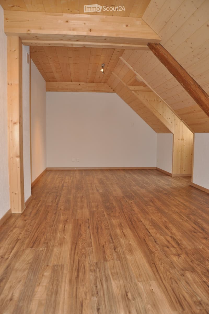 Empty attic space with wooden flooring, white walls, and exposed wooden beams on the ceiling. A wooden staircase leads upwards.