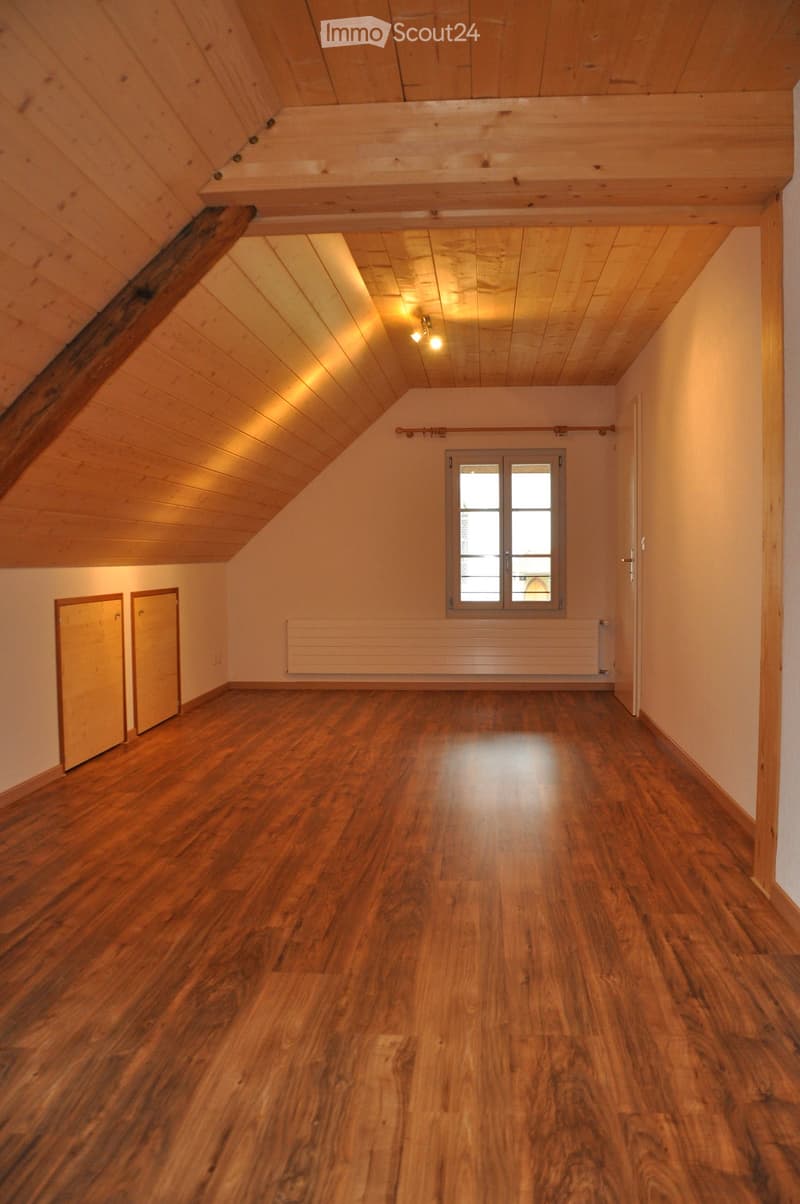 Empty room with wooden flooring, wooden ceiling beams, and natural light from a window. The space features a radiator and two doors, with no furniture or personal items present.