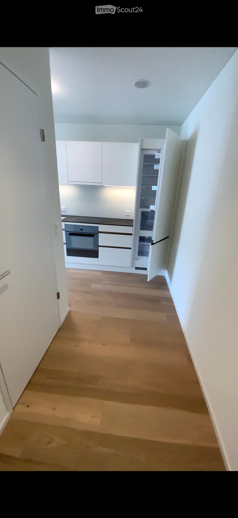 Empty hallway with wooden flooring leading to a modern kitchen area featuring white cabinets, a built-in oven, and a refrigerator. The kitchen is clean and unfurnished.