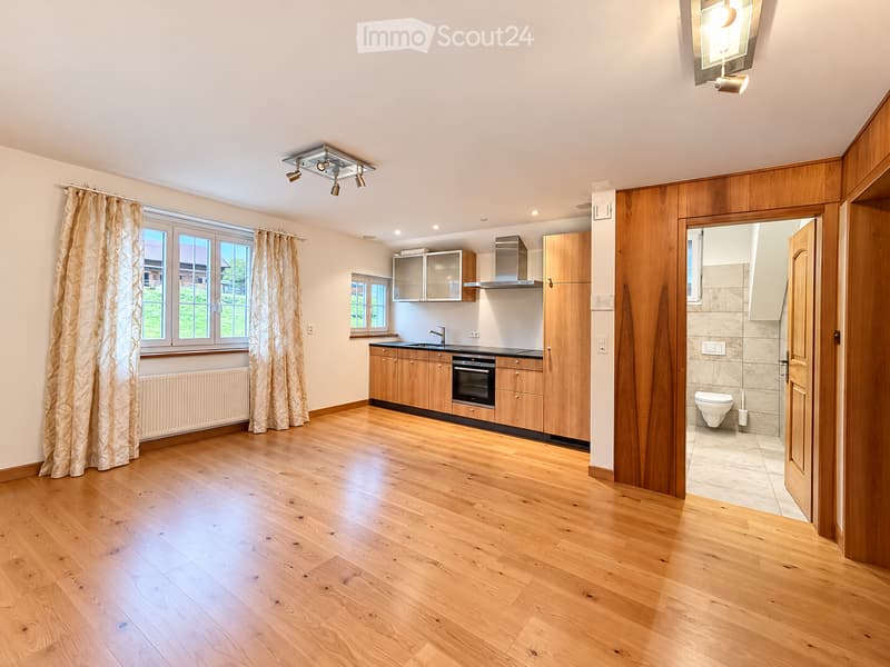 Bright, modern kitchen with wooden cabinets, stainless steel appliances, and a large window with curtains. The floor is made of light wooden planks, and a small bathroom is visible through an open doorway.