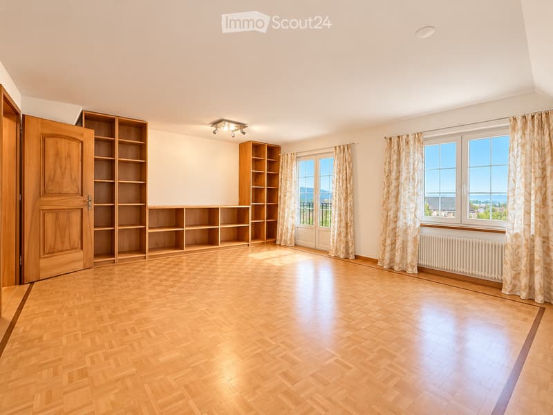 Empty room with wooden herringbone flooring, built-in wooden shelving units, large windows with curtains, and a radiator under one window.