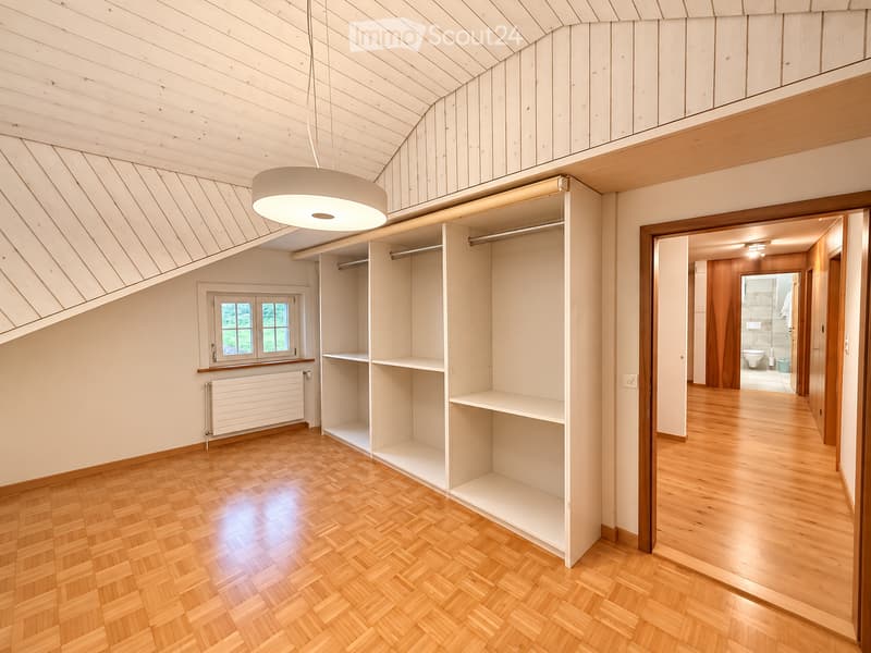 Empty room with wooden flooring, white walls, built-in closet with empty shelves, and a window with a view of greenery. The room features a modern pendant light fixture and a radiator under the window.