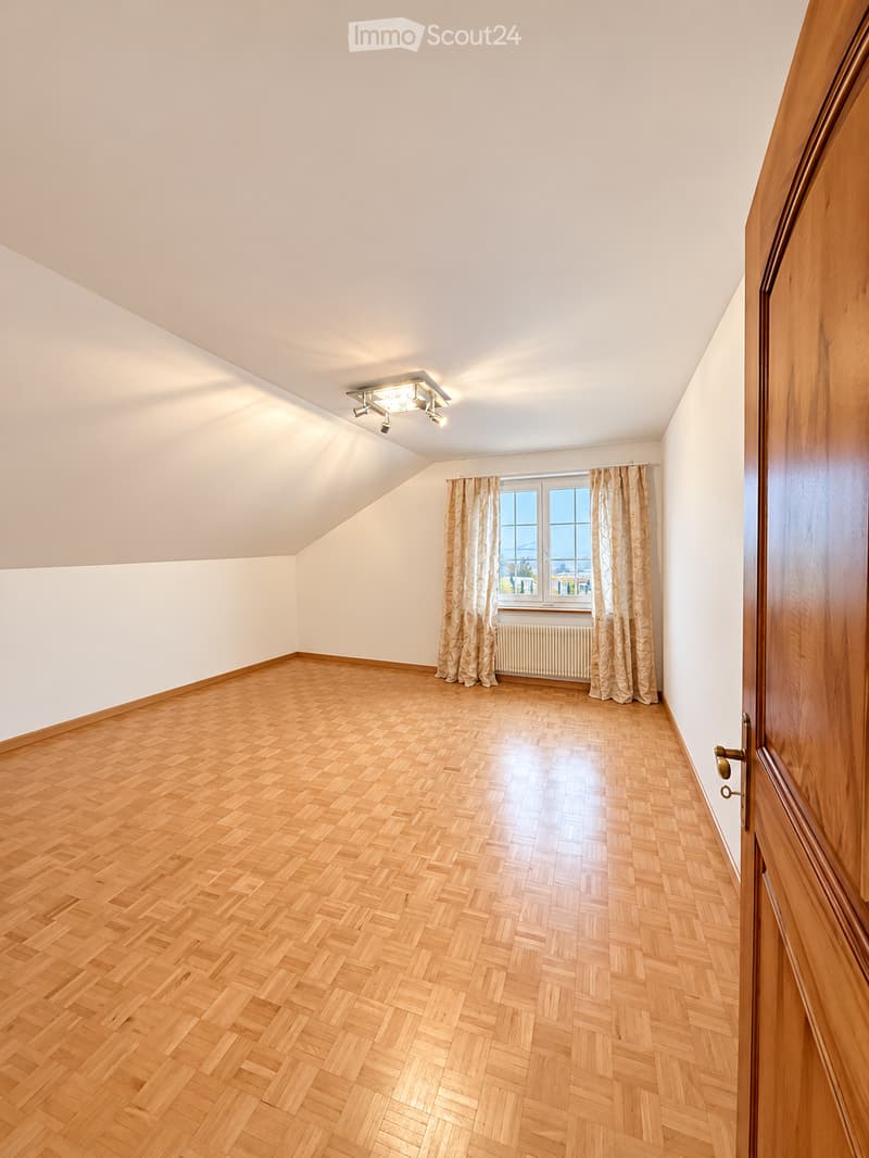 Empty room with wooden herringbone flooring, white walls, and a large window with curtains. The room features a slanted ceiling and a ceiling light fixture.