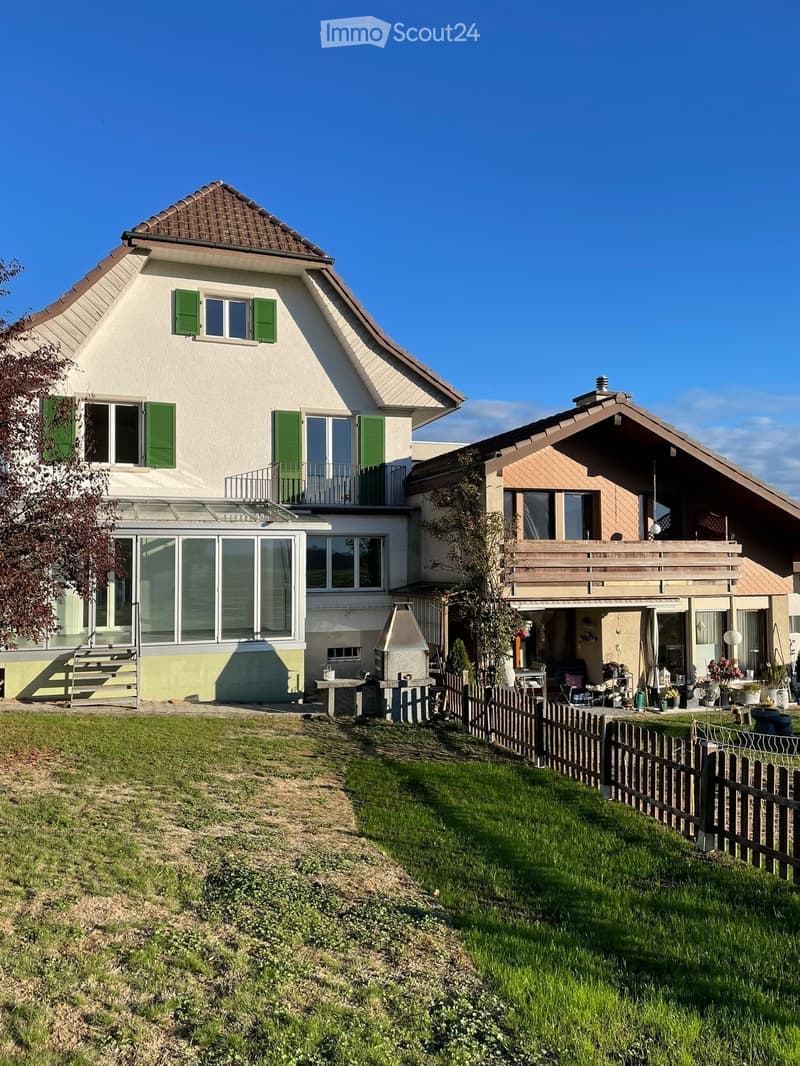 Two-story house with green shutters, a balcony, and a well-maintained garden with a wooden fence and outdoor seating area.