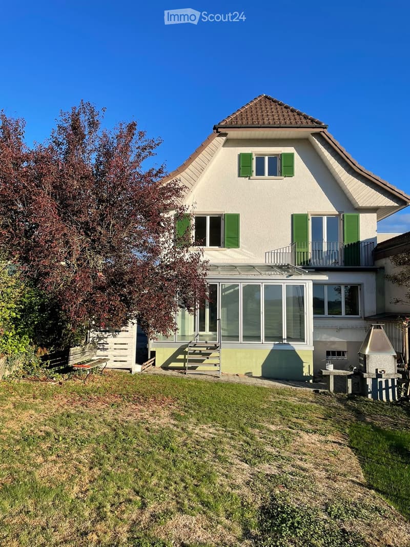Two-story house with white walls, green shutters, and a tiled roof. Features a balcony on the upper level and a patio area with a barbecue and seating.