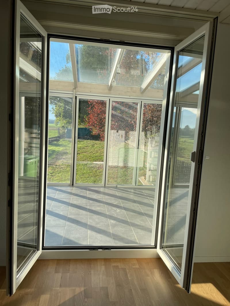 Bright, empty room with wooden flooring and large glass doors leading to a patio area with tiled flooring and outdoor view.