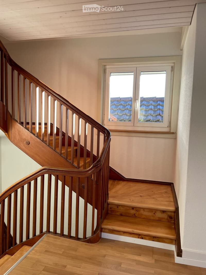 Spiral staircase with wooden steps and handrail, light-colored walls, and a window overlooking a roof.