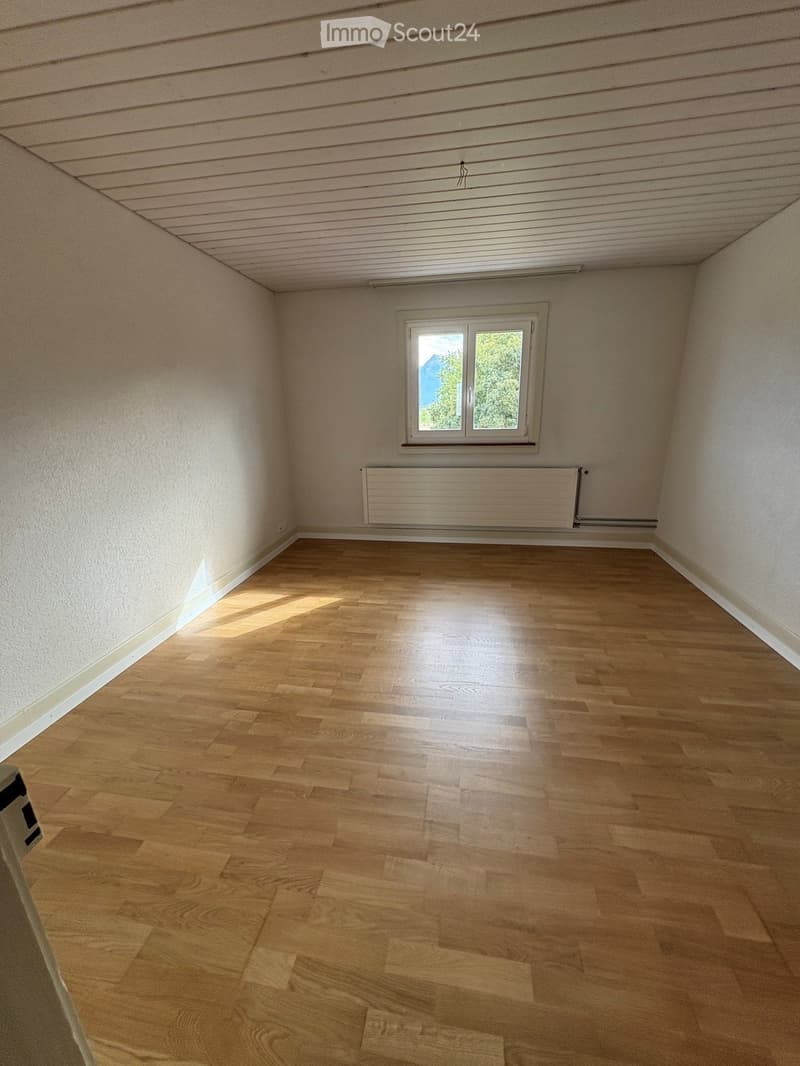 Empty room with wooden flooring, white walls, and a single window letting in natural light. A radiator is mounted on the wall below the window.