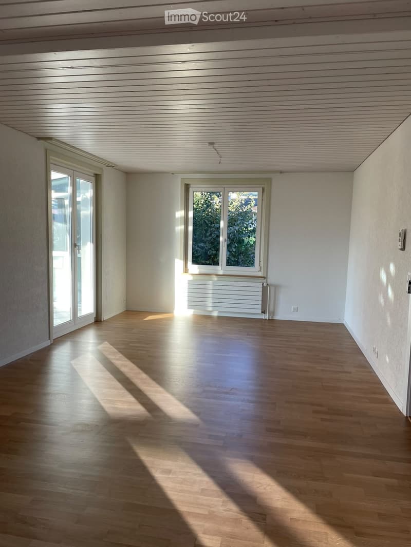 Empty room with wooden flooring, white walls, and large windows allowing natural light to enter. The space is unfurnished and features a radiator below one of the windows.