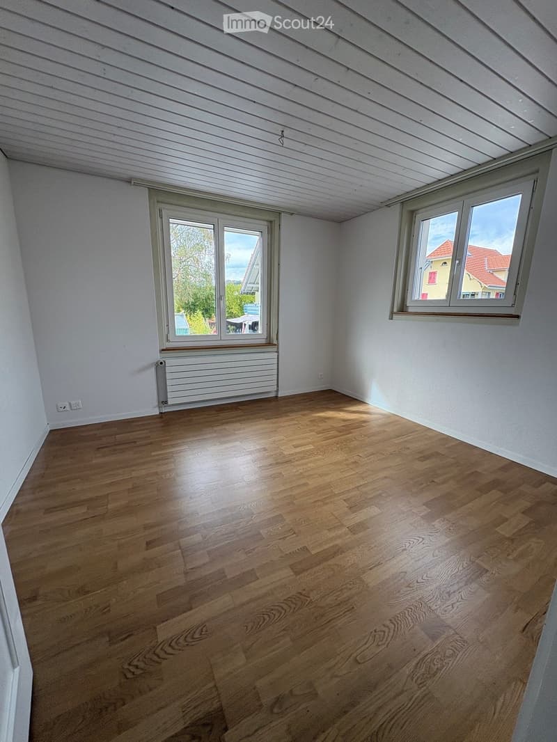 Empty room with wooden flooring, white walls, and large windows offering a view of the outside. The room features a radiator and electrical outlets.