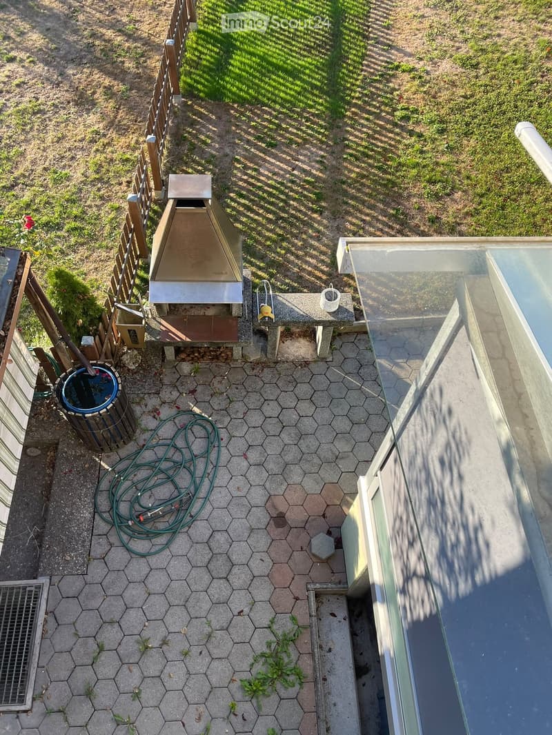 A small patio area with hexagonal tiles, a wooden fence, a small shed, a hose, a trash bin, and a small pool with a glass cover.