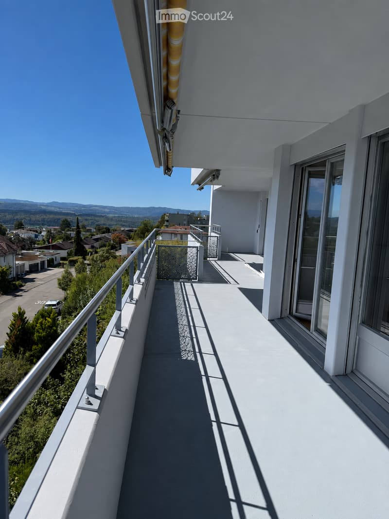 Balcony with white flooring, metal railing, and a view of a suburban neighborhood with trees and houses.