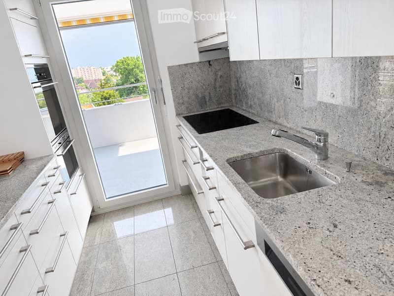 Modern kitchen with white cabinets, granite countertops, stainless steel sink, and built-in oven. Large window offers a view of greenery and buildings.