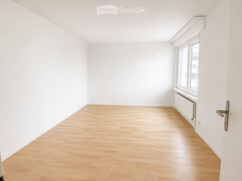 Empty room with white walls, wooden flooring, and a large window with a radiator below it.