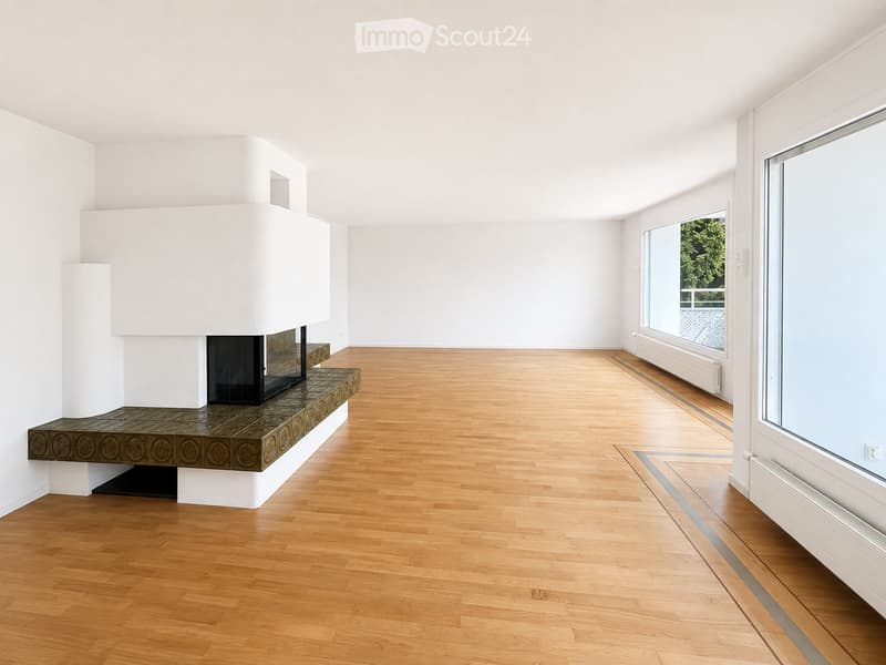 Empty room with light wooden flooring, white walls, and a built-in fireplace with a decorative tiled surround. Large windows provide natural light.