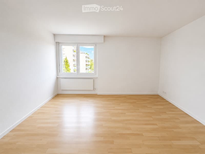 Empty room with light wooden flooring, white walls, and a large window with a view of trees and buildings outside.