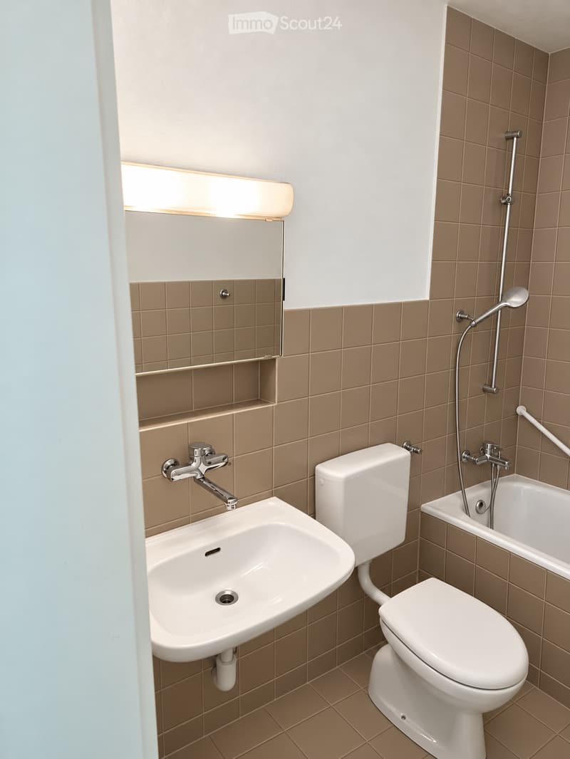 Modern bathroom with beige tiles, a white sink, toilet, bathtub, and a mirror with built-in lighting.