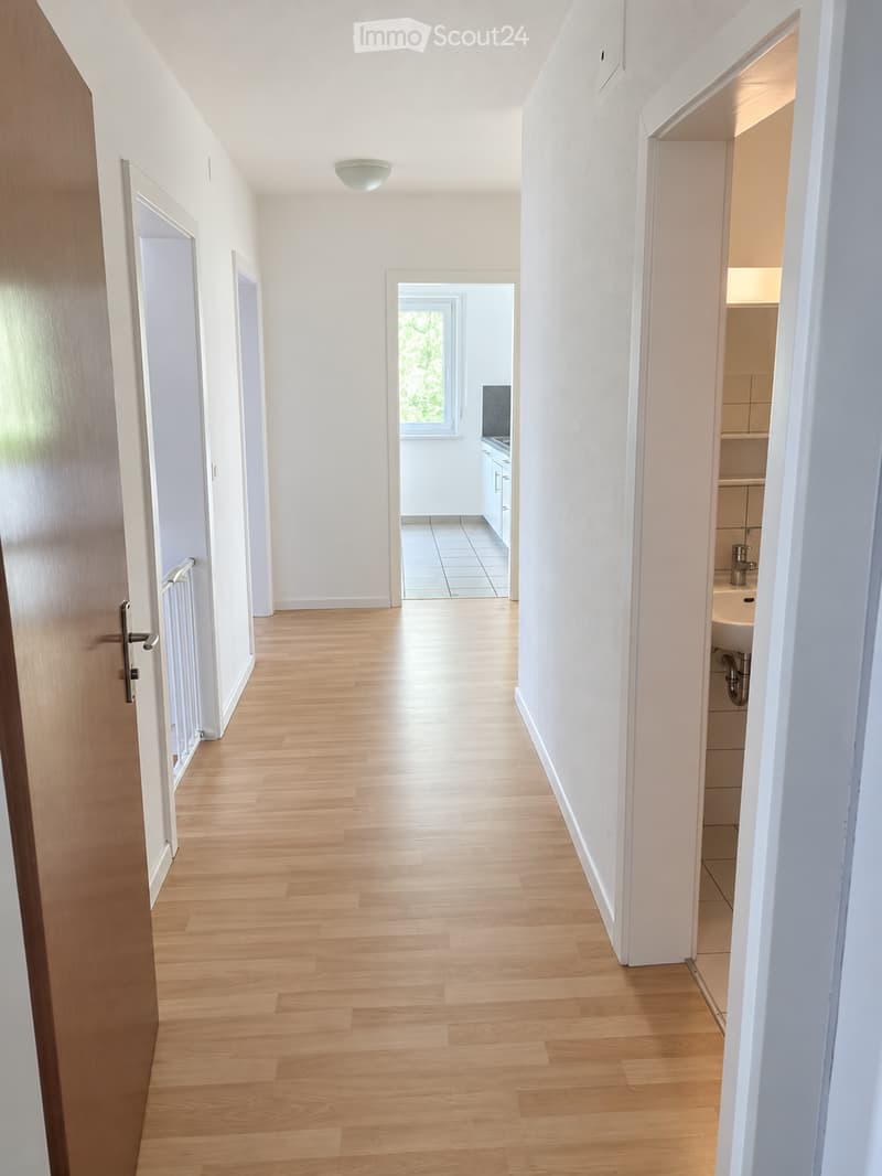 Bright hallway with wooden flooring, white walls, and several doors leading to different rooms. Natural light streams in from a window at the end of the corridor.