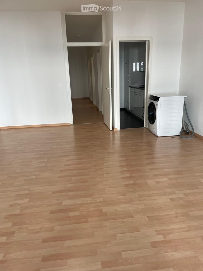 Empty room with light wooden flooring, white walls, and a doorway leading to another room. A washing machine is visible in an adjacent area.