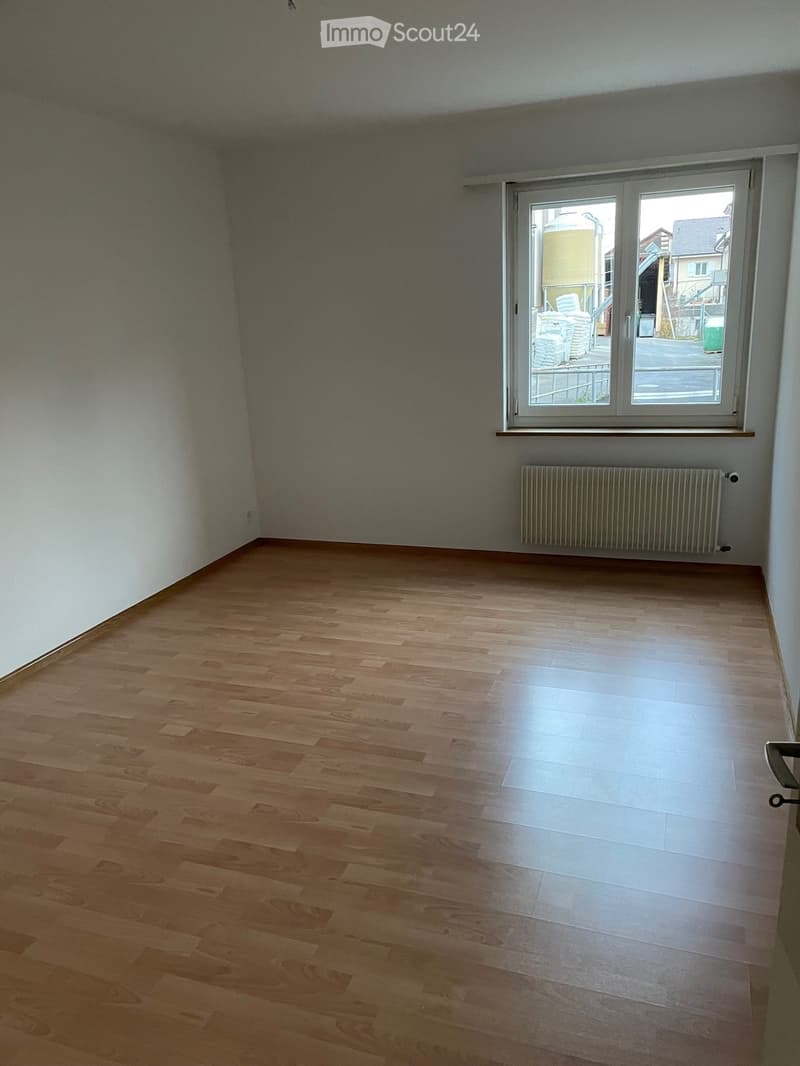 Empty room with wooden flooring, white walls, a window with a view of the street, and a radiator below the window.