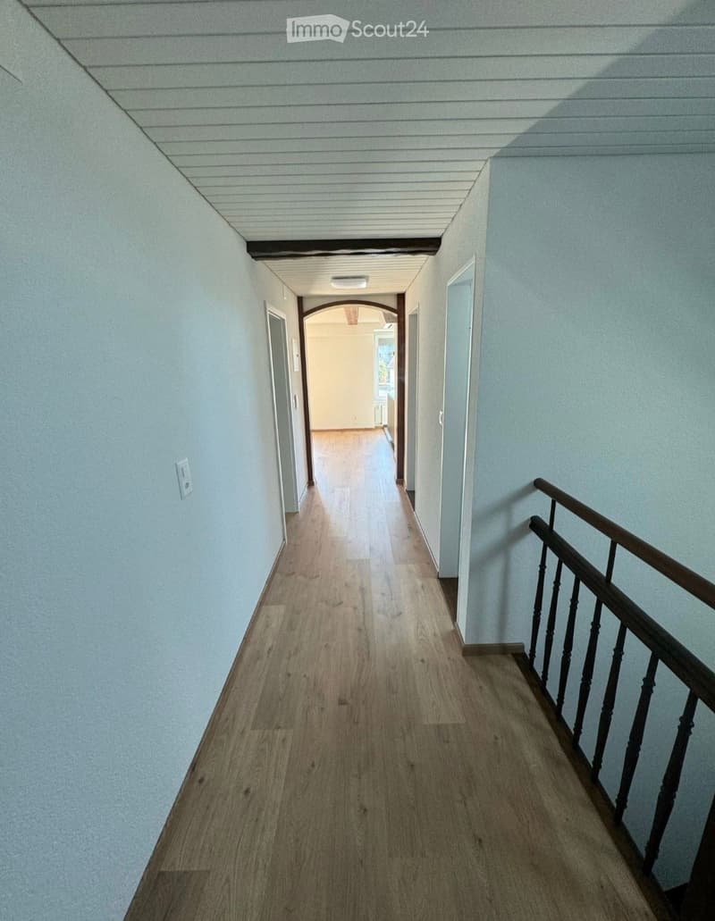 A clean, well-lit hallway with light wooden flooring, white walls, and a wooden railing on one side. The ceiling has a simple, paneled design, and there are several doors leading to other rooms.