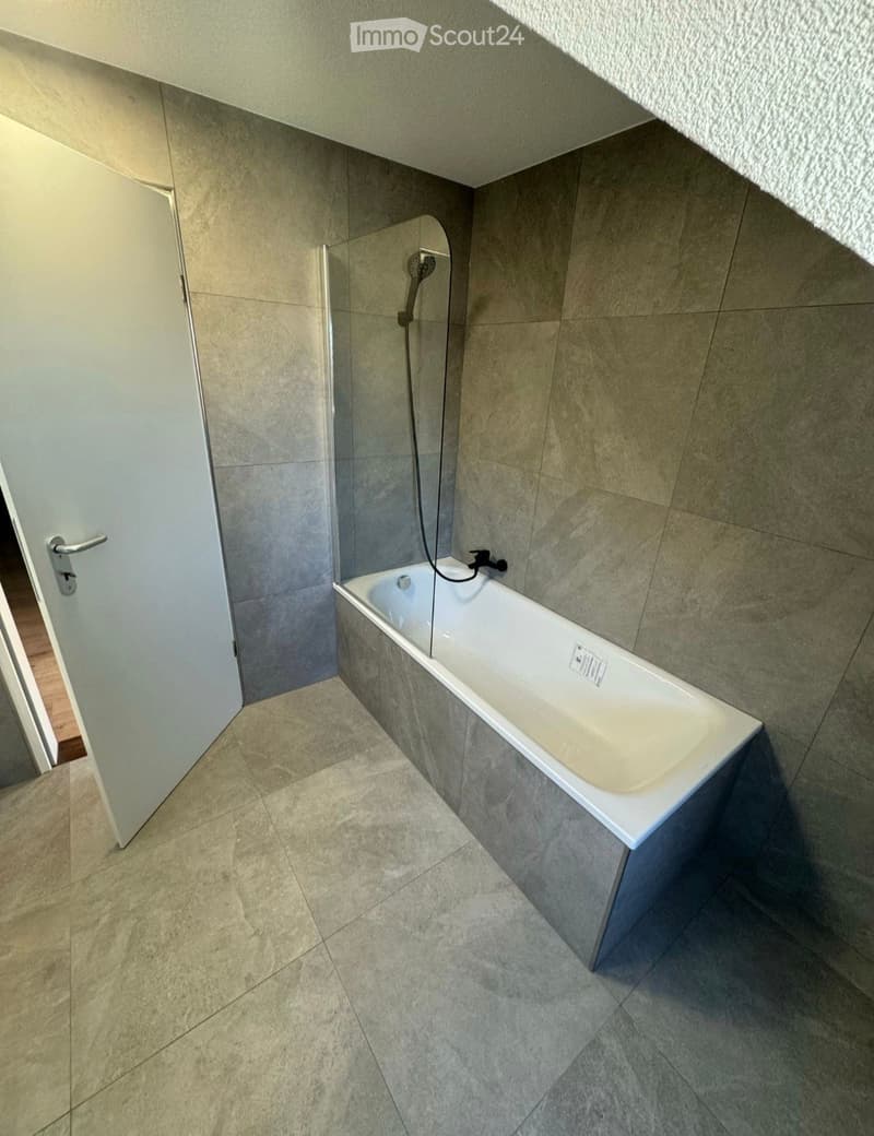 Modern bathroom with a white bathtub, glass shower enclosure, tiled walls and floor, and a white door.