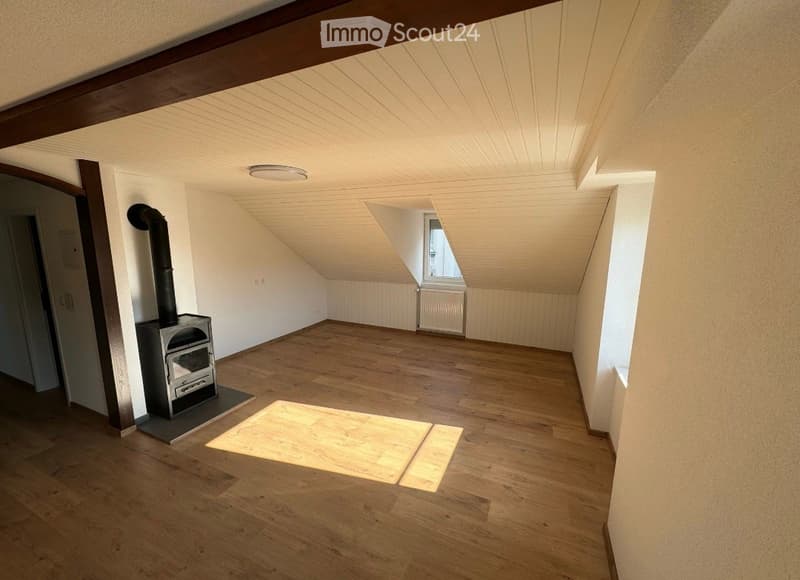 Empty attic room with wooden flooring, exposed wooden beams, and a small wood-burning stove. Natural light streams in from a dormer window.