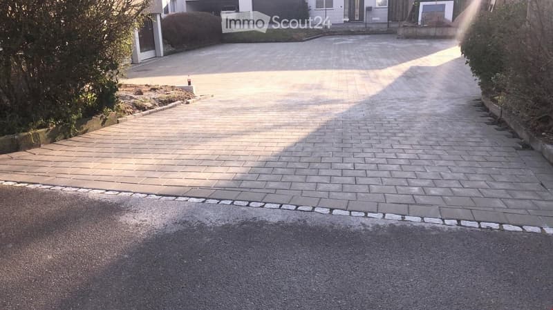 A paved driveway leading to a house, bordered by low hedges and a small garden area on one side.