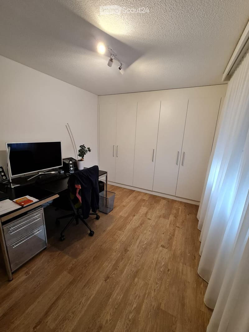 A modern, well-lit room with wooden flooring, white walls, and built-in white cabinets. A desk with a computer monitor, keyboard, and chair is positioned near a window with a curtain. A small potted plant sits on the desk, and a jacket is draped over the