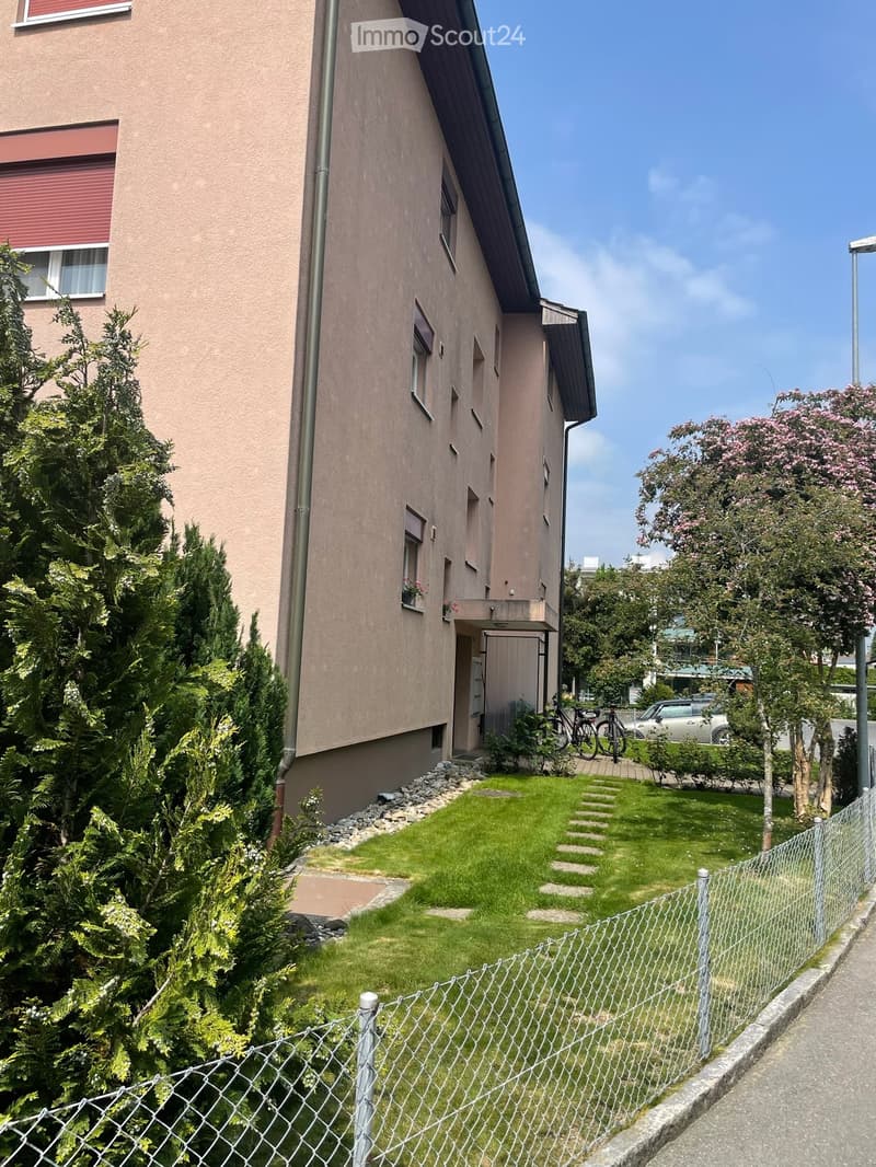 A well-maintained garden area with a small lawn, stone pathway, and a chain-link fence. The garden is bordered by a house with a balcony and several trees, including conifers and flowering shrubs.