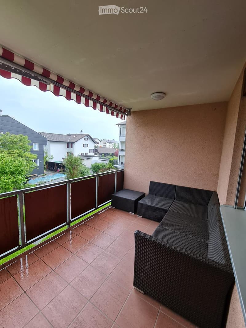 Balcony with tiled floor, wicker sofa, and a small stool, featuring a view of neighboring houses and greenery.