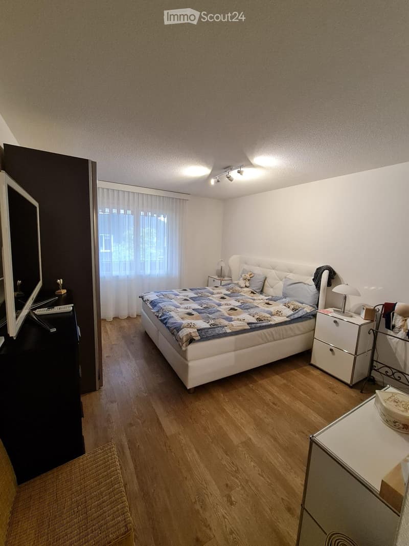 Cozy bedroom with wooden flooring, a large bed with a patterned quilt, nightstands with lamps, a mirror, and a wicker chair near the door.