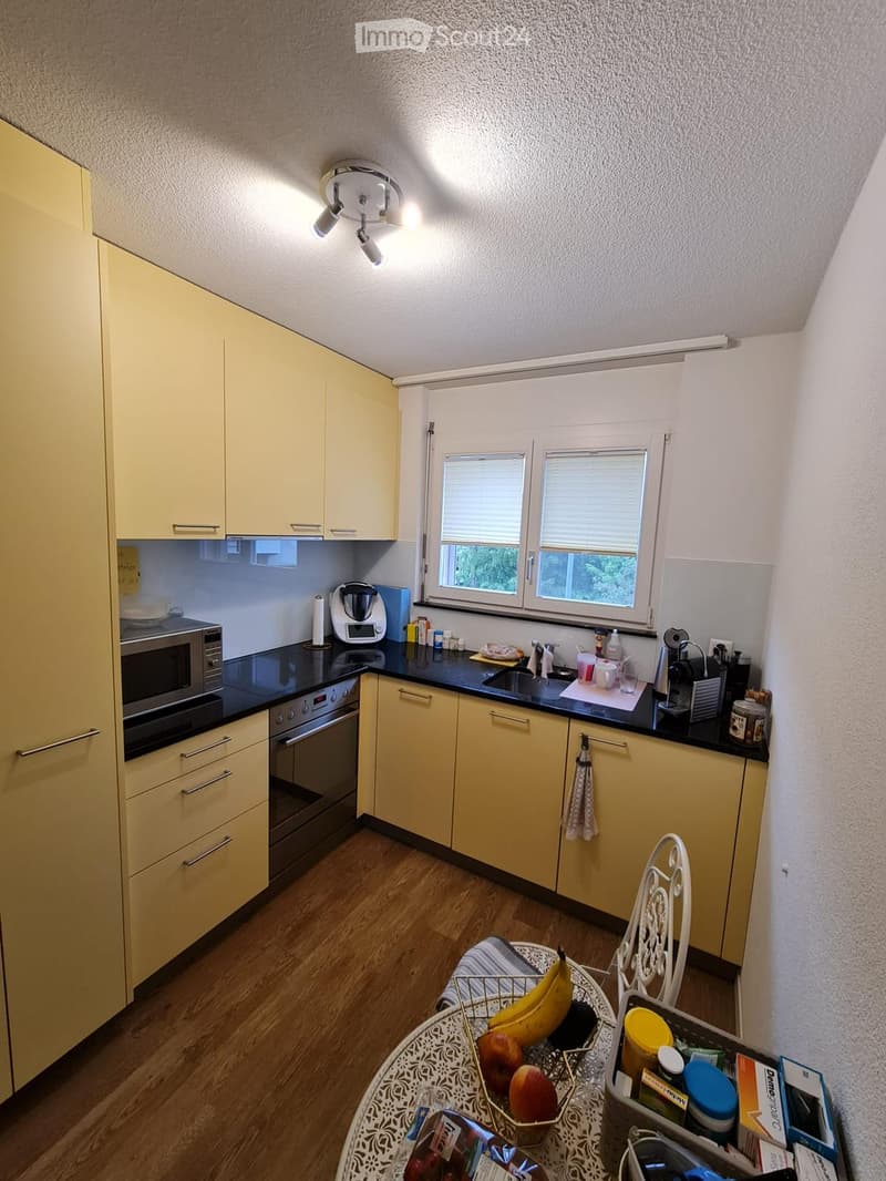 Bright kitchen with light yellow cabinets, black countertops, and wooden flooring. Features a round table with a fruit basket, a microwave, oven, and various kitchen appliances.