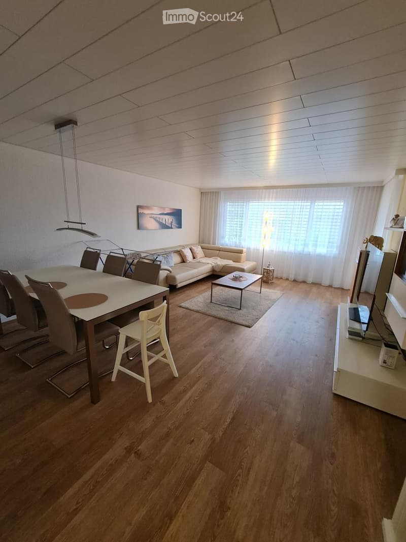 Modern living and dining area with light wood flooring, a cream-colored sectional sofa, a glass coffee table, a dining table with chairs, and a television on a stand.
