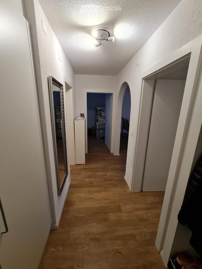 A narrow hallway with wooden flooring, white walls, and a ceiling light fixture. There are two open doors leading to other rooms, and a mirror is mounted on the left wall.