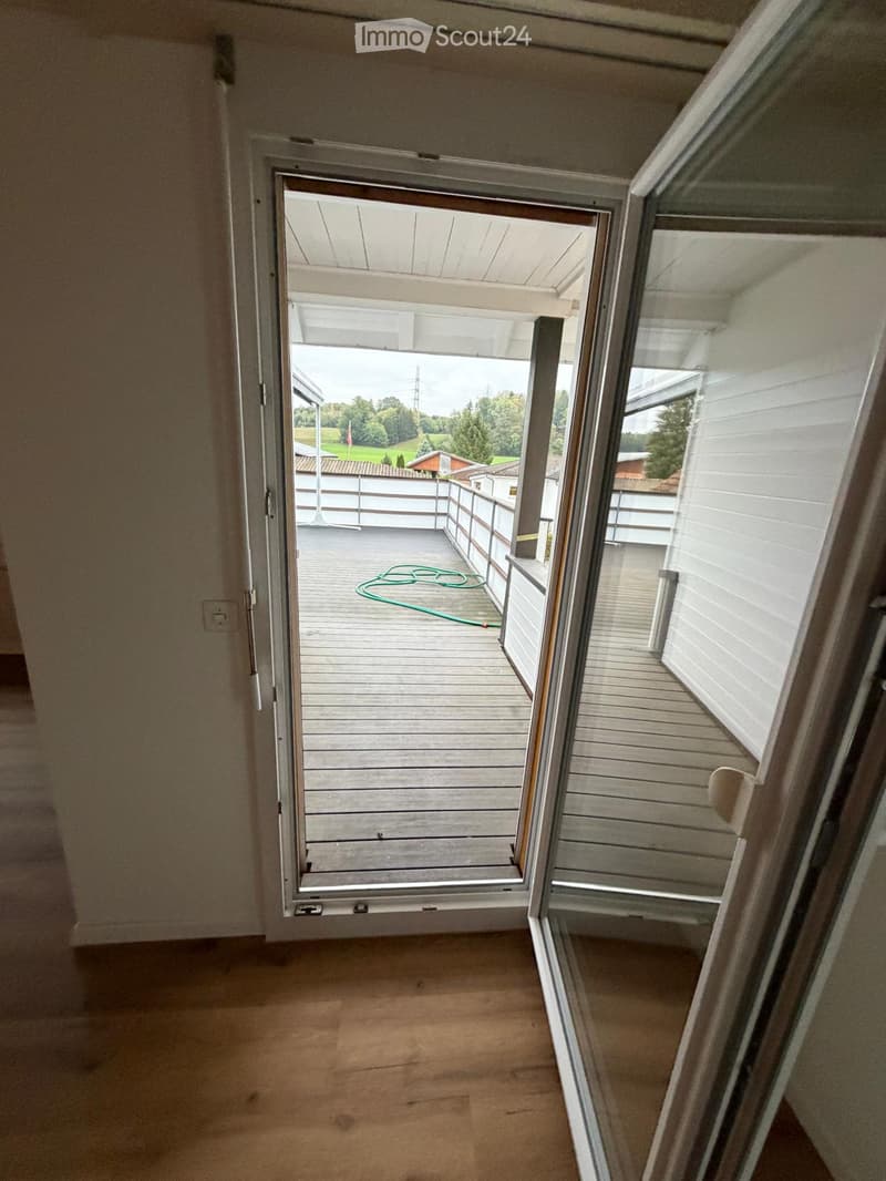 Open sliding door leads to a wooden deck with a railing, a green hose on the floor, and a view of a suburban landscape with trees and houses.