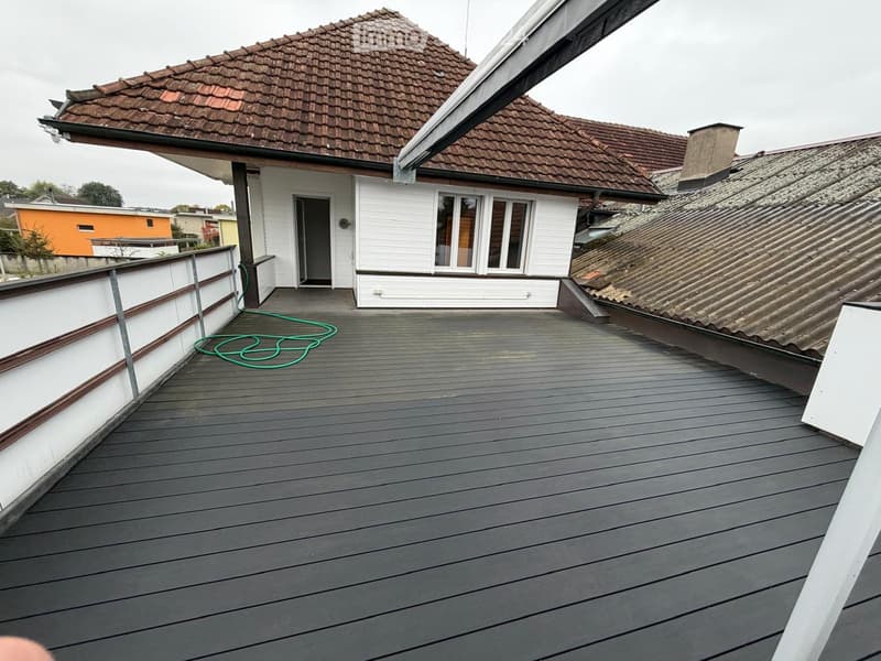Roof terrace with wooden decking, white railing, and a green garden hose coiled on the floor. Adjacent house with white walls, large windows, and a tiled roof is visible.