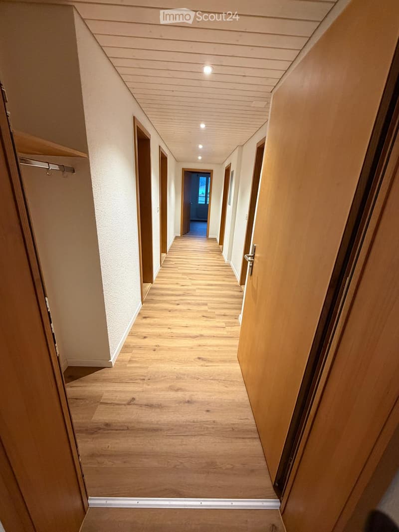 A well-lit hallway with wooden flooring, white walls, and several doors on both sides. The ceiling has recessed lighting, and the hallway appears clean and organized.