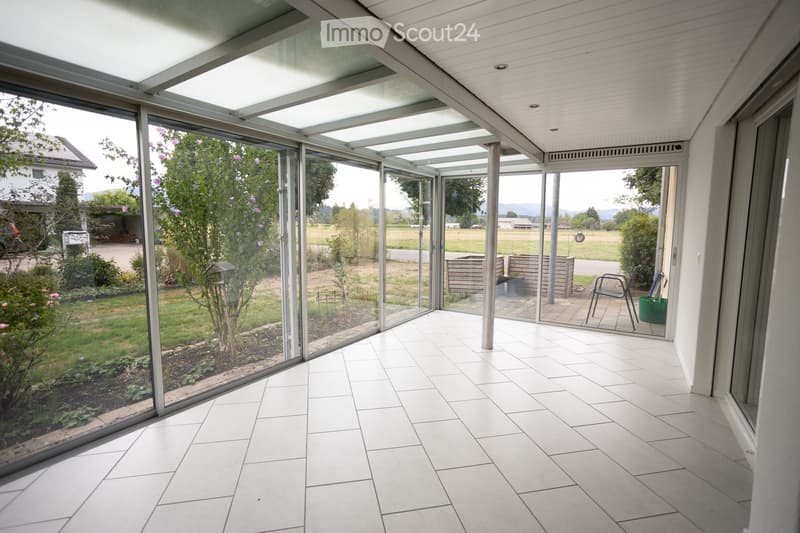 Bright, enclosed sunroom with large glass walls and a tiled floor, offering views of a garden and open landscape.