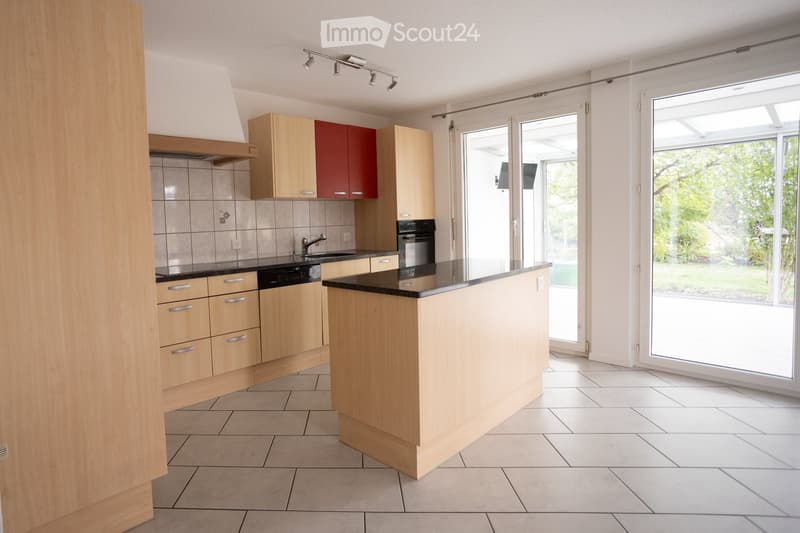 Modern kitchen with light wooden cabinets, a central island, and large windows overlooking a garden.
