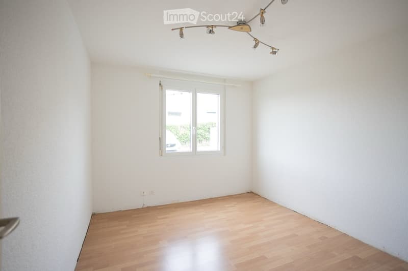 Empty room with light wooden flooring, white walls, and a large window with blinds. The room features a modern track light fixture on the ceiling.