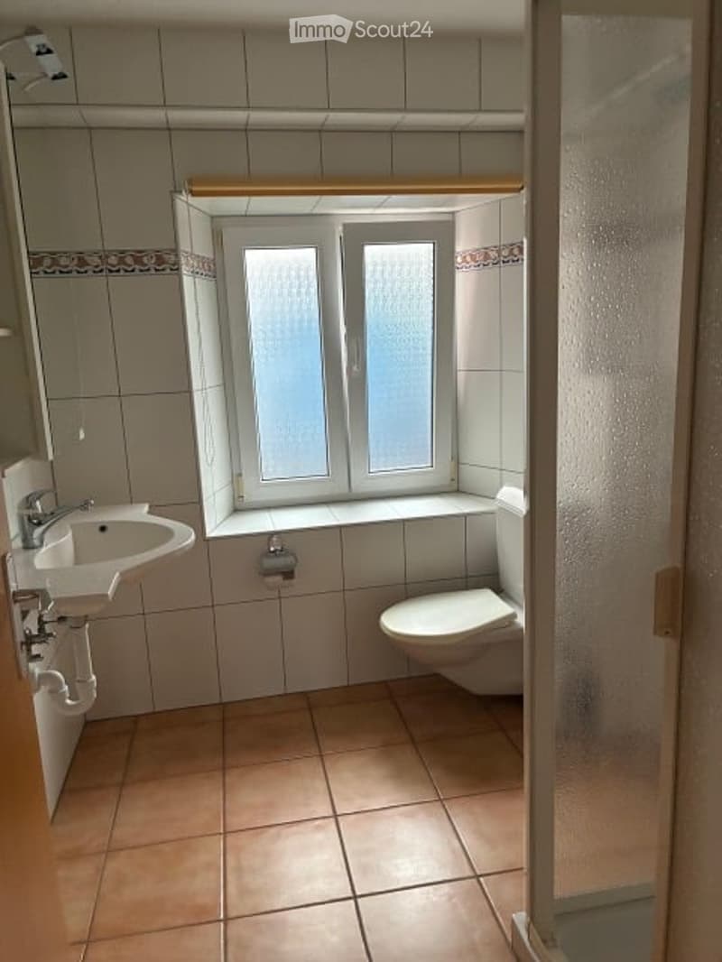 Bathroom with beige tiled walls and floor, a white sink with a chrome faucet, a toilet, and a frosted glass shower enclosure. A window with frosted glass panes is above the toilet.