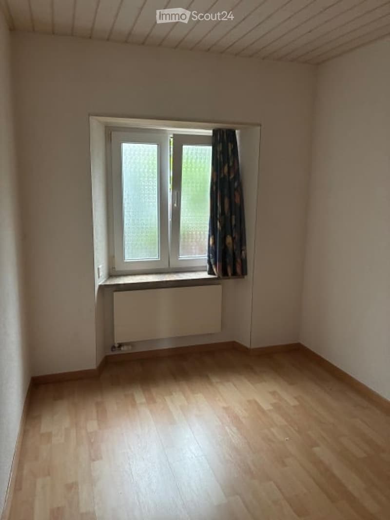 Empty room with wooden flooring, white walls, a window with a curtain, and a radiator below the window.