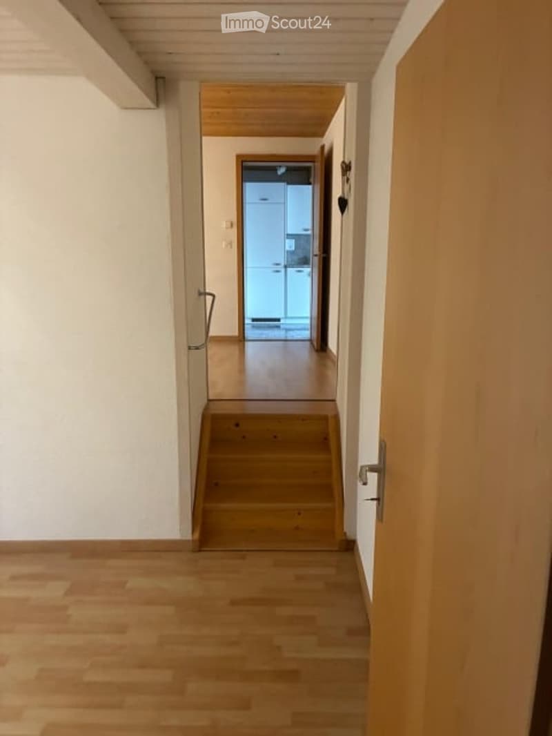 A narrow hallway with wooden flooring and white walls, leading to a staircase and an open doorway to another room.