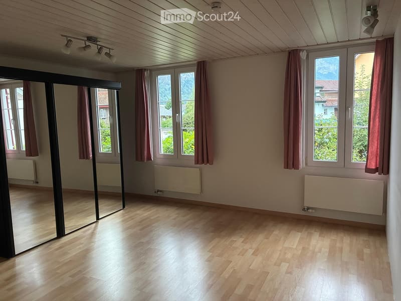 Empty room with wooden flooring, large windows with pink curtains, radiators, and a large mirror on one wall.
