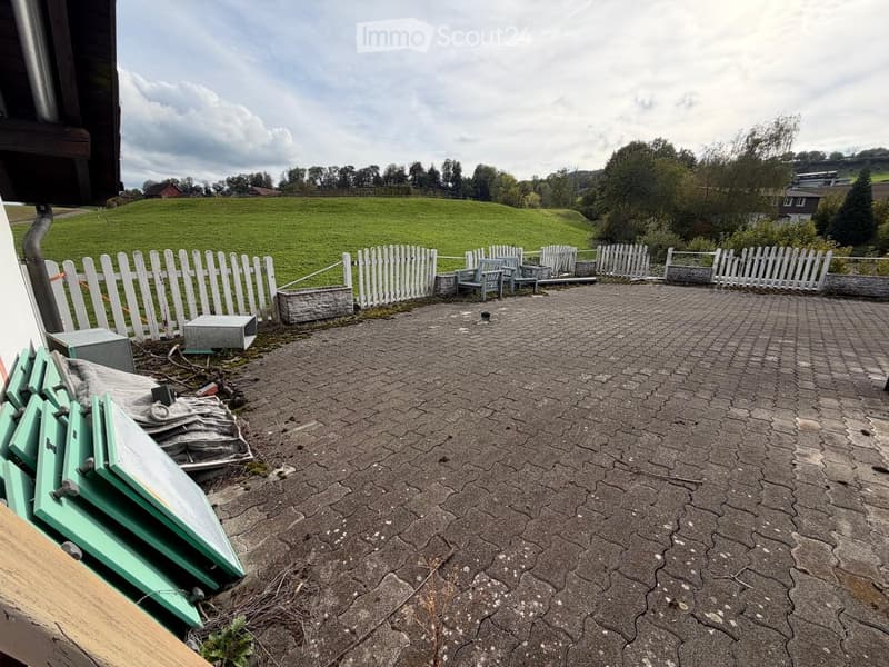 Outdoor patio area with cracked concrete surface, white picket fence, and scattered outdoor furniture including chairs and tables.