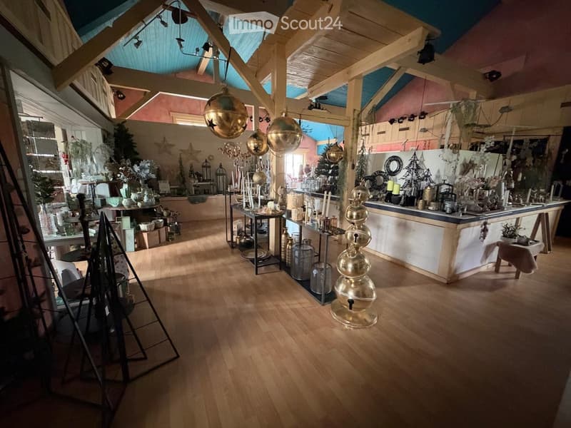 A spacious, well-lit interior with wooden flooring, exposed wooden beams, and a high, slanted ceiling. The room features decorative items such as vases, candles, mirrors, and shelves displaying various objects. There is a small table and a stool near the